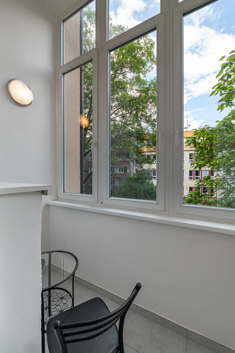 A sunroom with a black chair and glass table, featuring large windows overlooking trees and nearby apartment buildings.