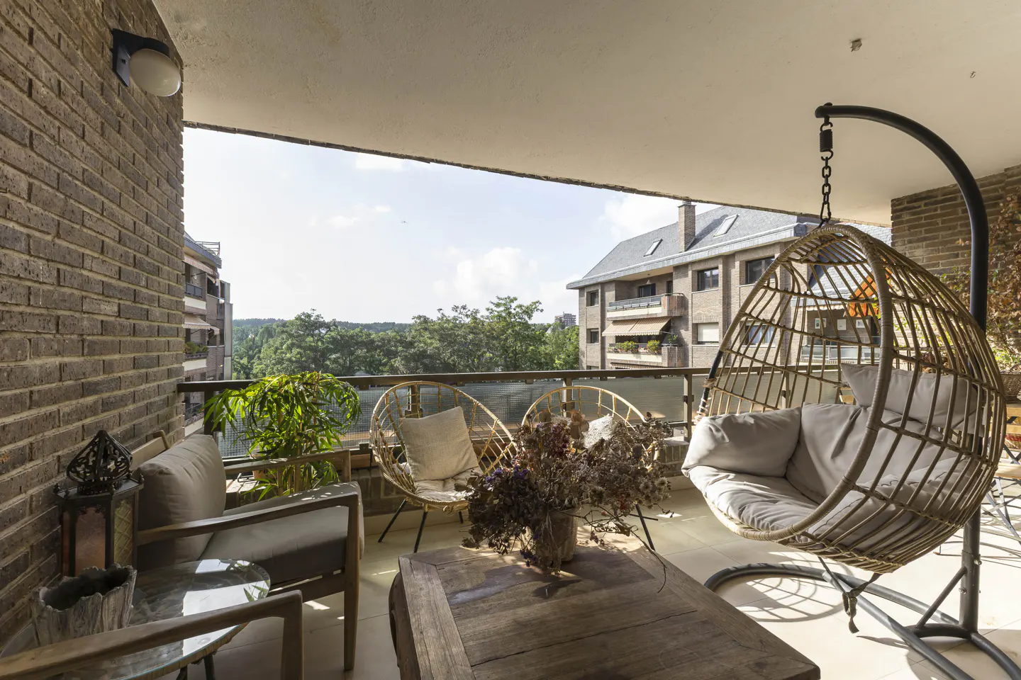 A furnished balcony with a hanging chair, table, and seating overlooks a green landscape.