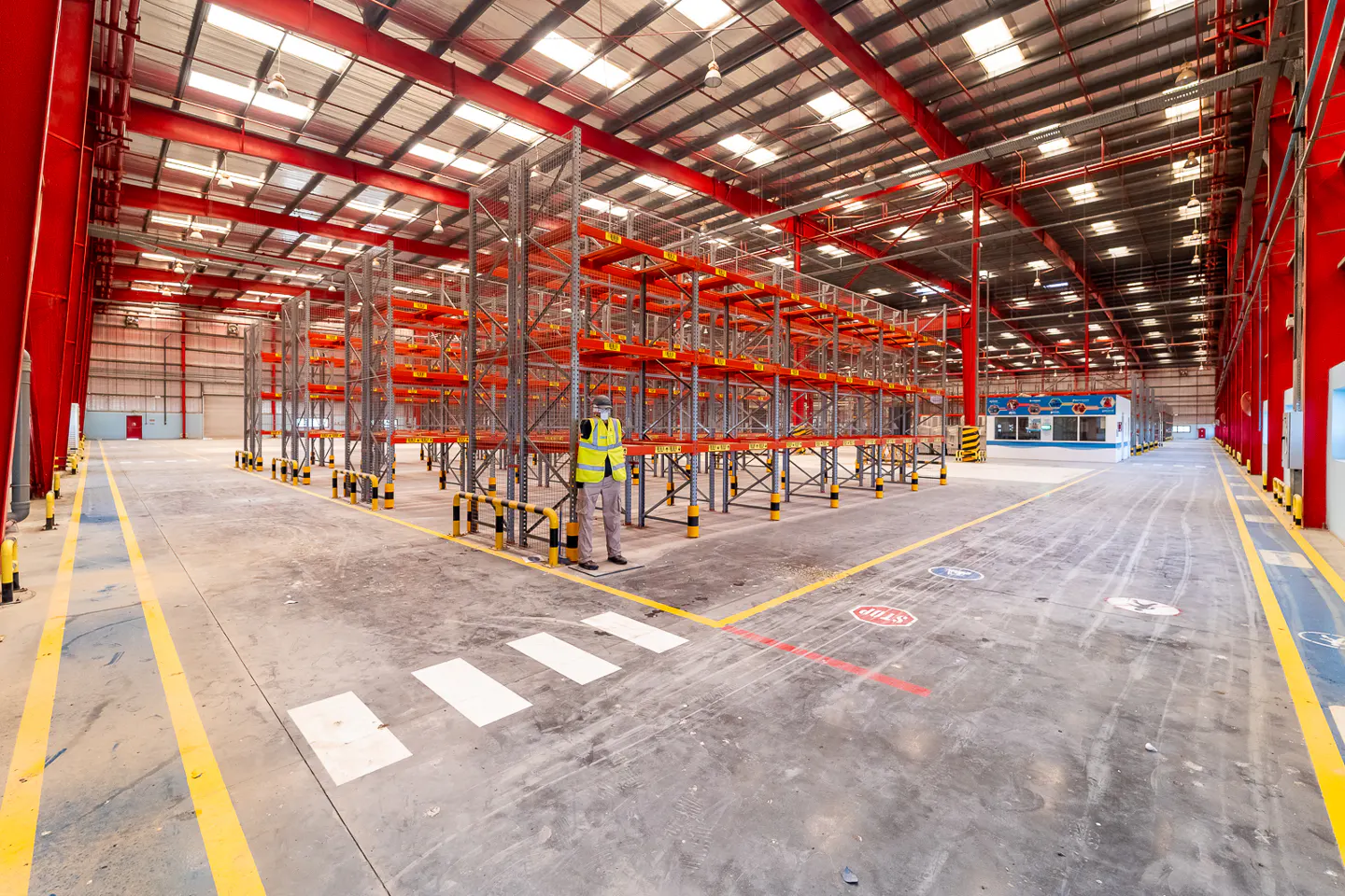 A large, empty warehouse with red support beams and metal shelving. A worker in a yellow vest stands near a crosswalk.