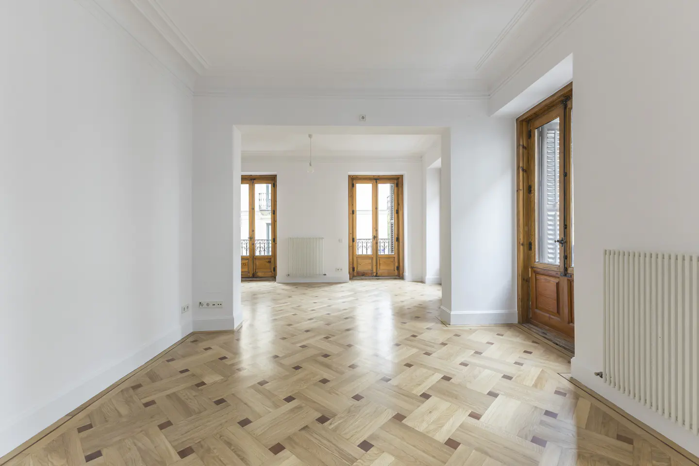 Empty room with white walls, herringbone wood floor, and wood-framed glass doors leading to balconies. Radiators are visible.