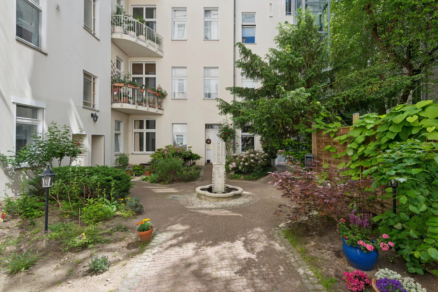 A quiet apartment courtyard with a stone fountain, lush greenery, potted flowers, and a paved walkway.