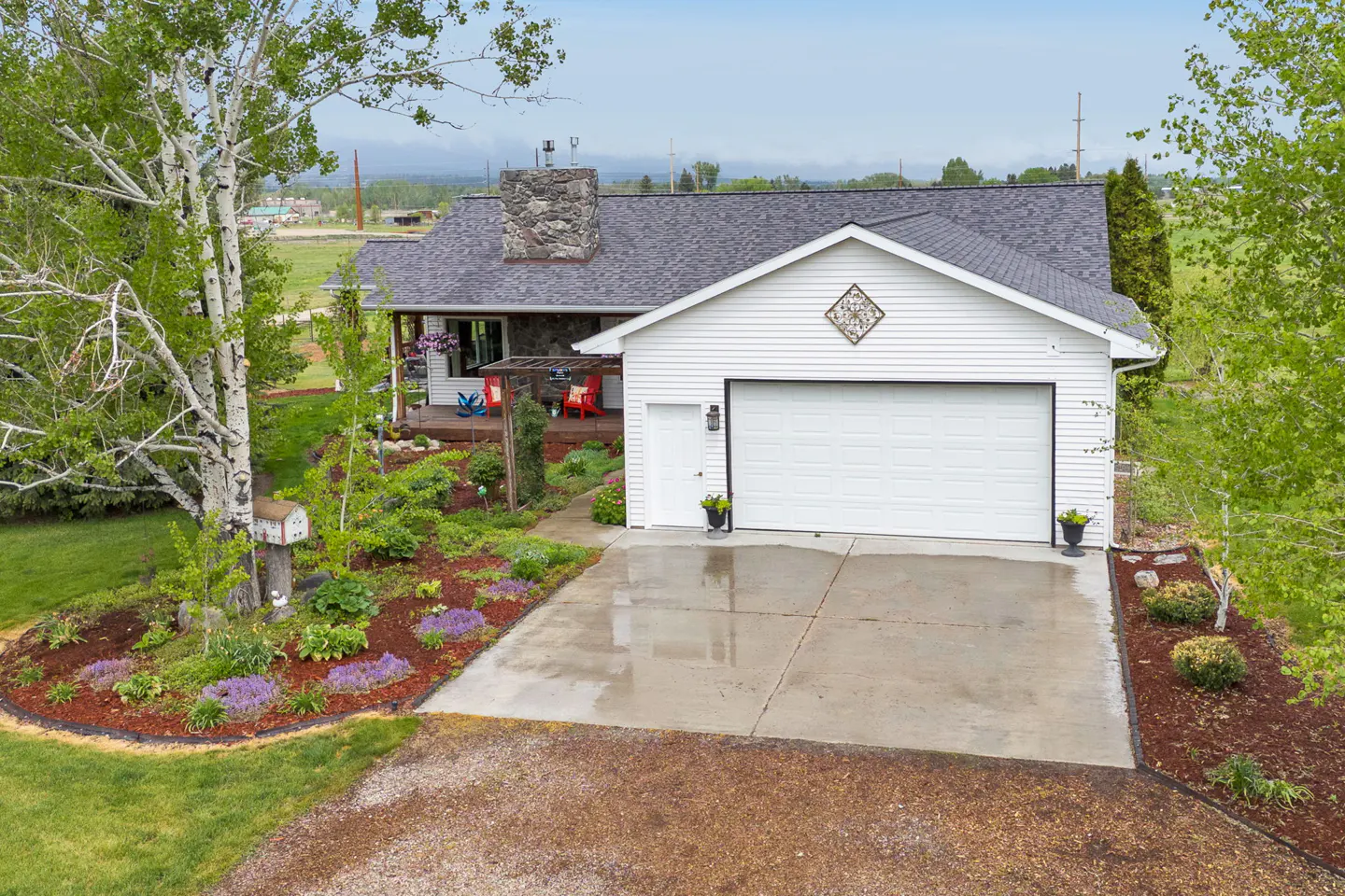 Aerial view of a white house with a gray roof, stone chimney, and a two-car garage. A garden with purple flowers lines the driveway.