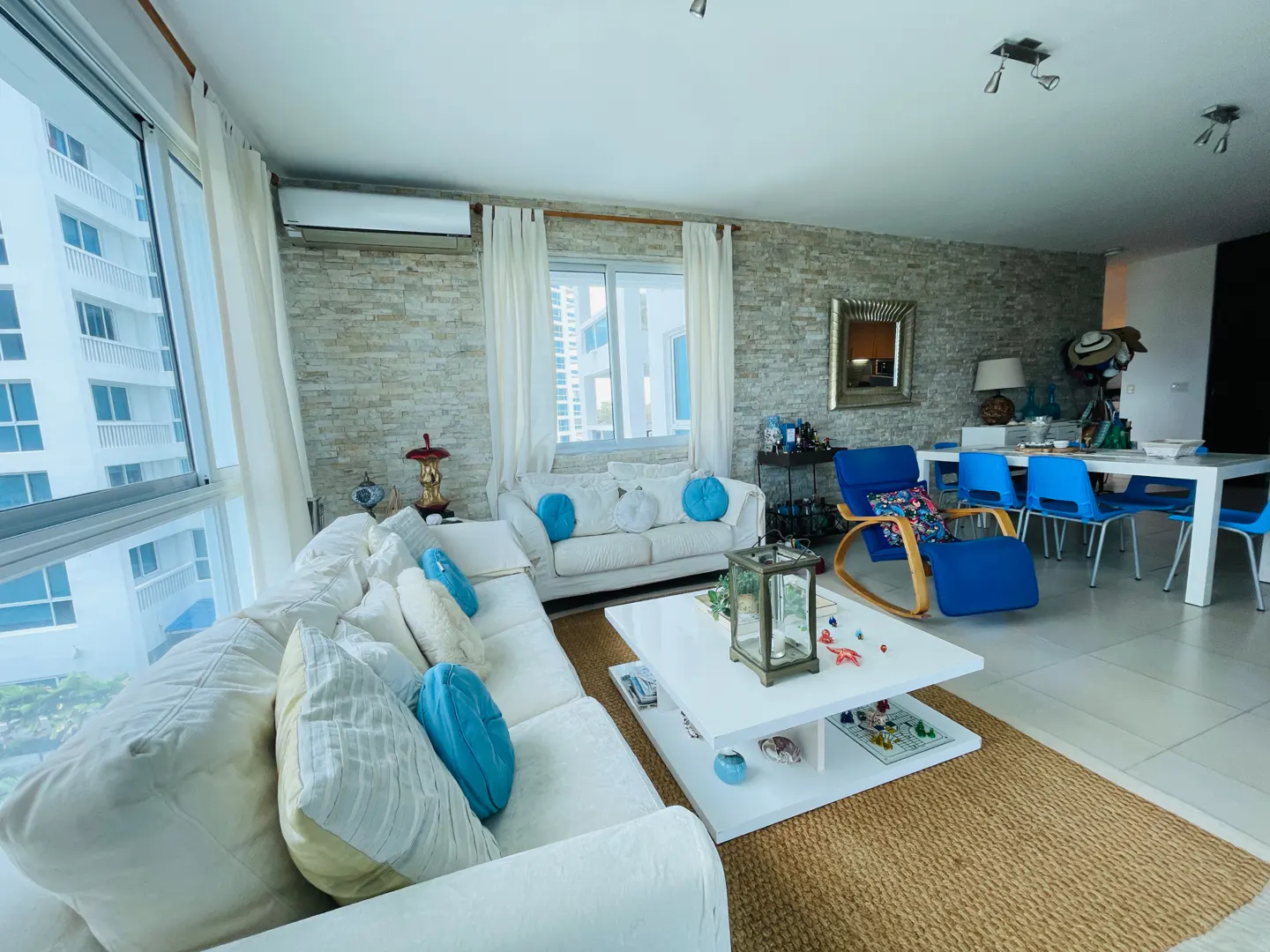 Bright living room with white sofas, blue pillows, and a white coffee table on a jute rug. A dining table with blue chairs sits in the background.