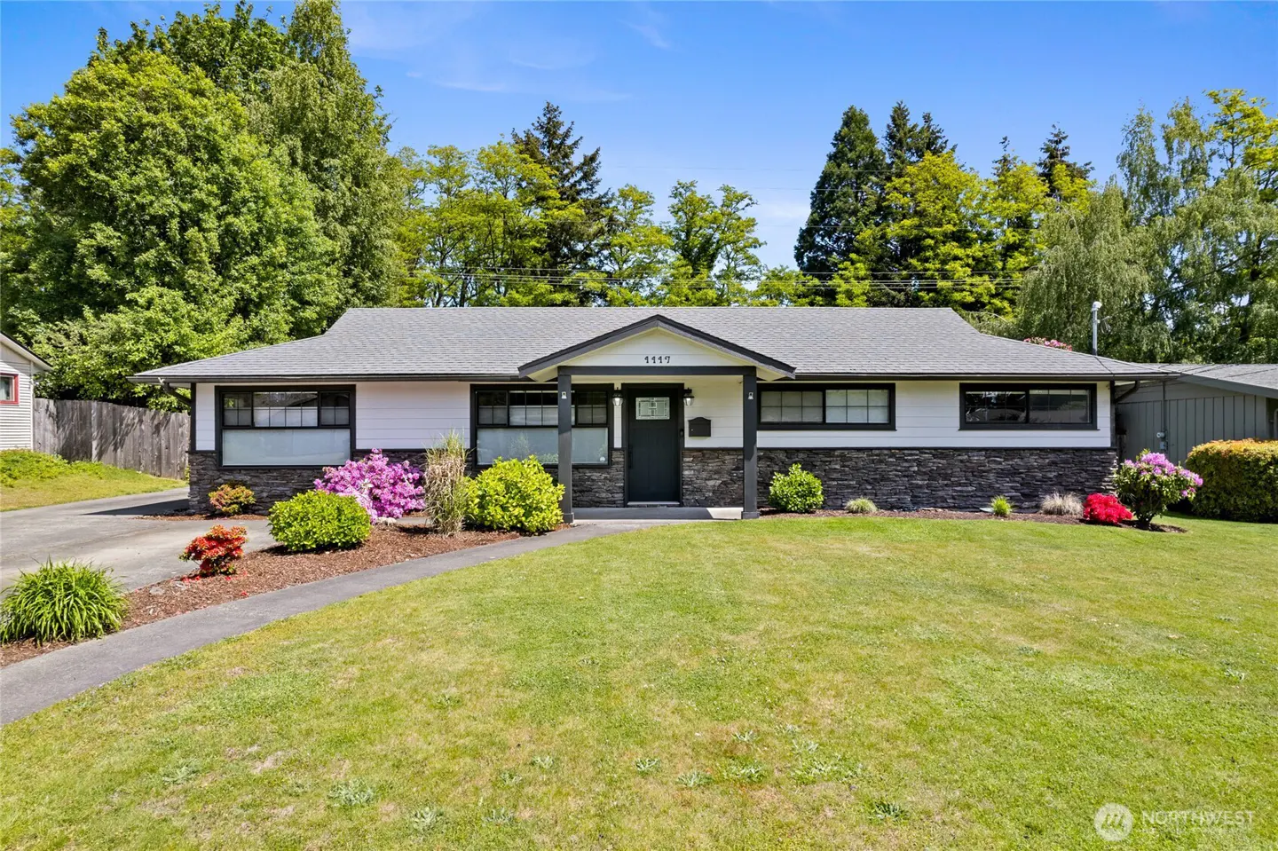 A single-story home with white siding, a gray roof, and black trim, surrounded by a green lawn and trees.