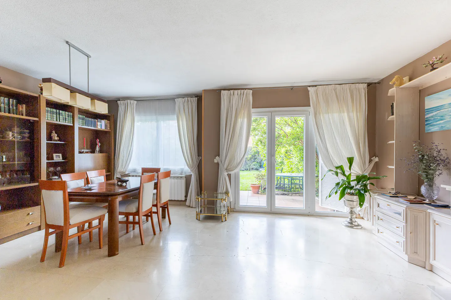 Bright dining room with a wood table, white chairs, and a view of a green garden through large glass doors. Built-in bookshelves and cabinets line the walls.