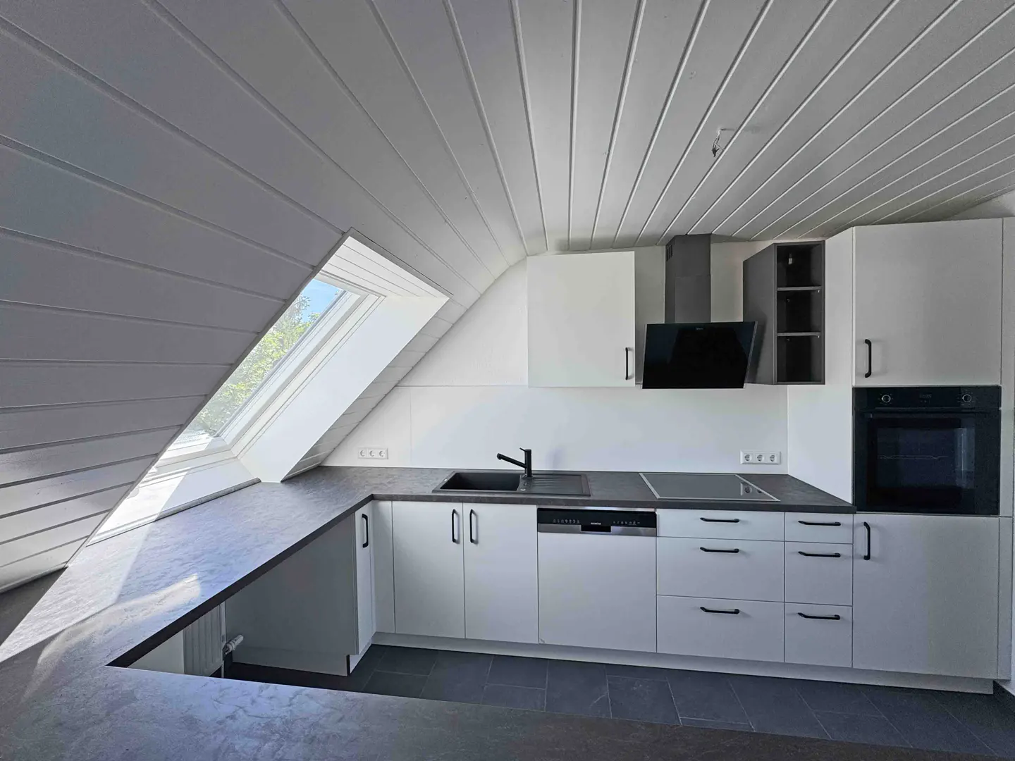 Modern attic kitchen with white cabinets, dark countertops, built-in oven, and a sloped ceiling with a skylight.
