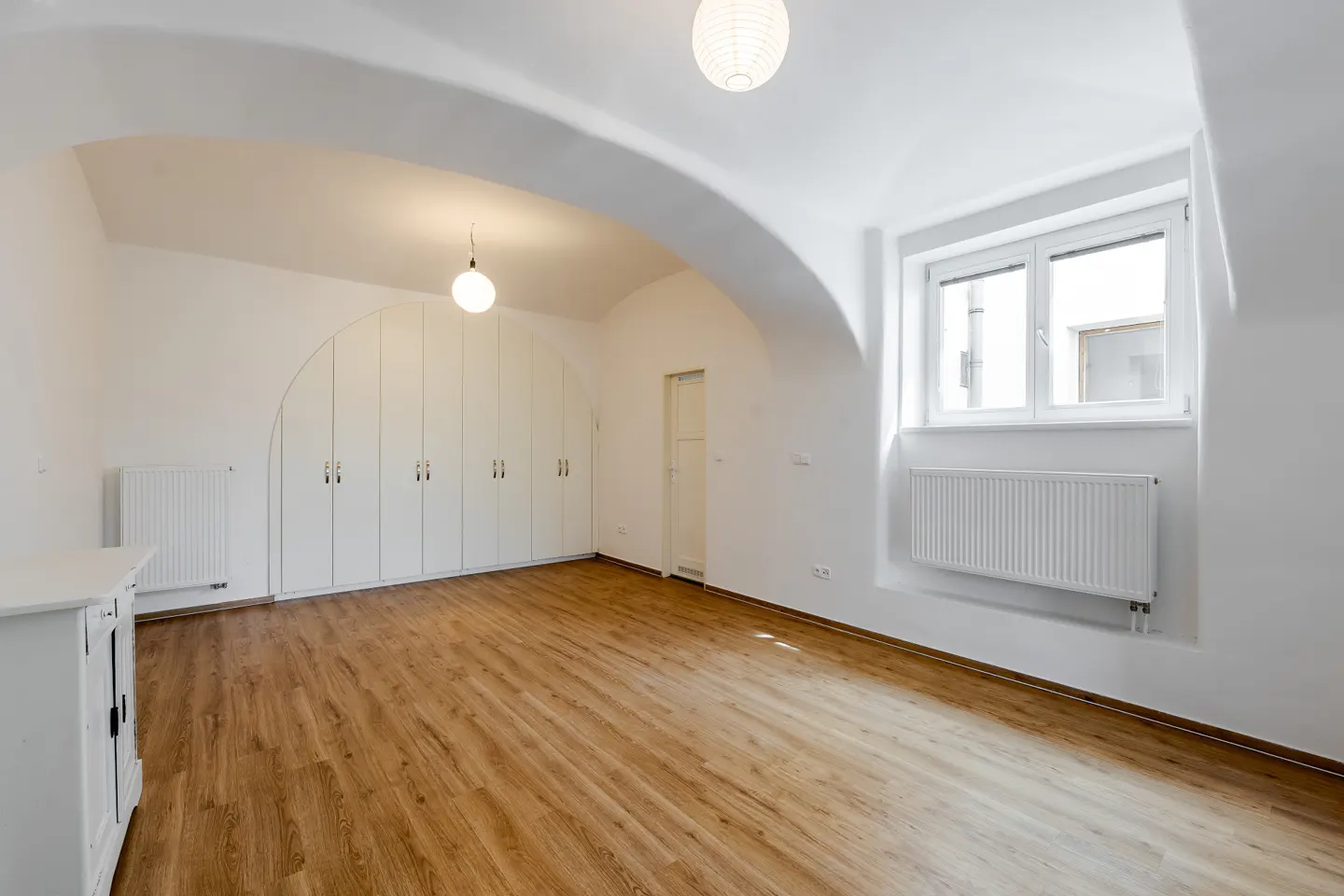 Bright, empty room with wood floors, white walls, and arched ceiling. A wall of white closets, a window, and a radiator are visible.
