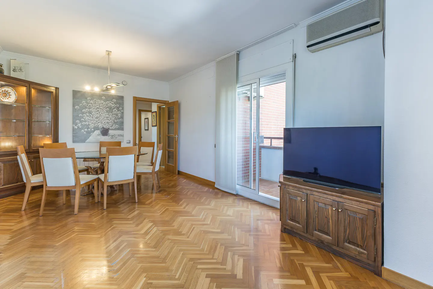 Bright living room with wood floors, a dining table with white chairs, and a TV on a wooden cabinet near a sliding glass door.