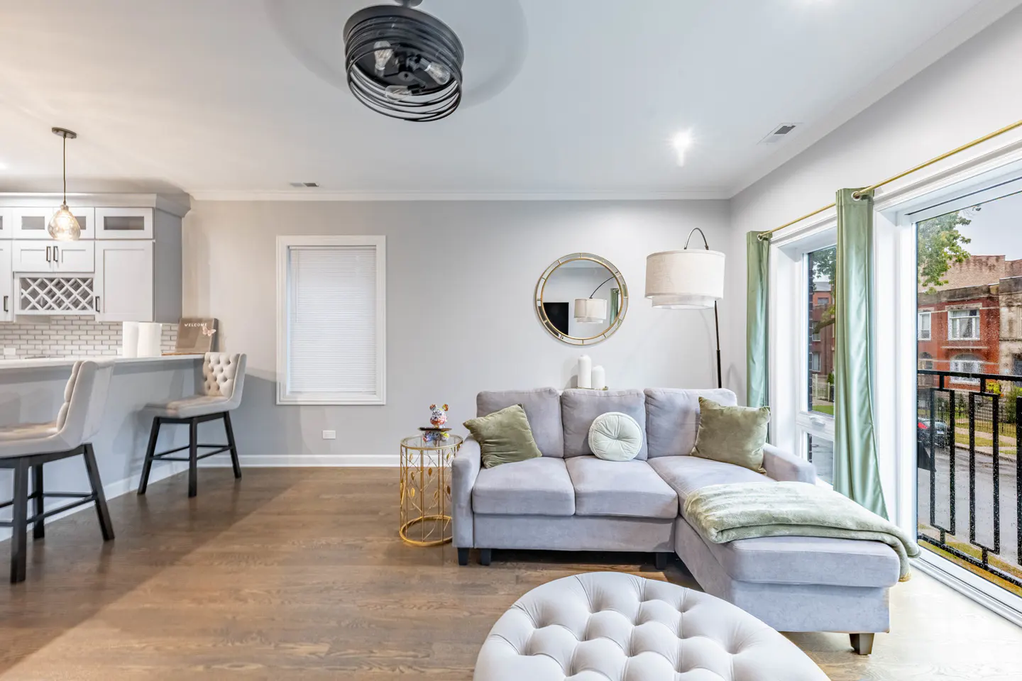 Living room with gray sectional sofa, round mirror, and light wood floors. Kitchen bar with stools visible in the background. Balcony with green curtains.
