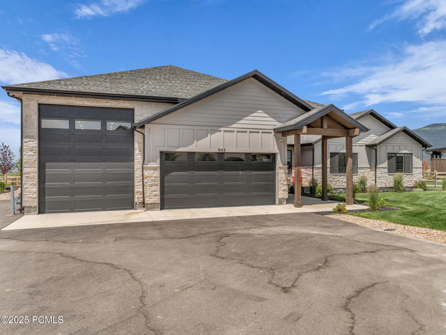 Exterior view of a modern, single-story home with gray siding, stone accents, and a dark gray roof under a blue sky. Two-car garage with dark doors.