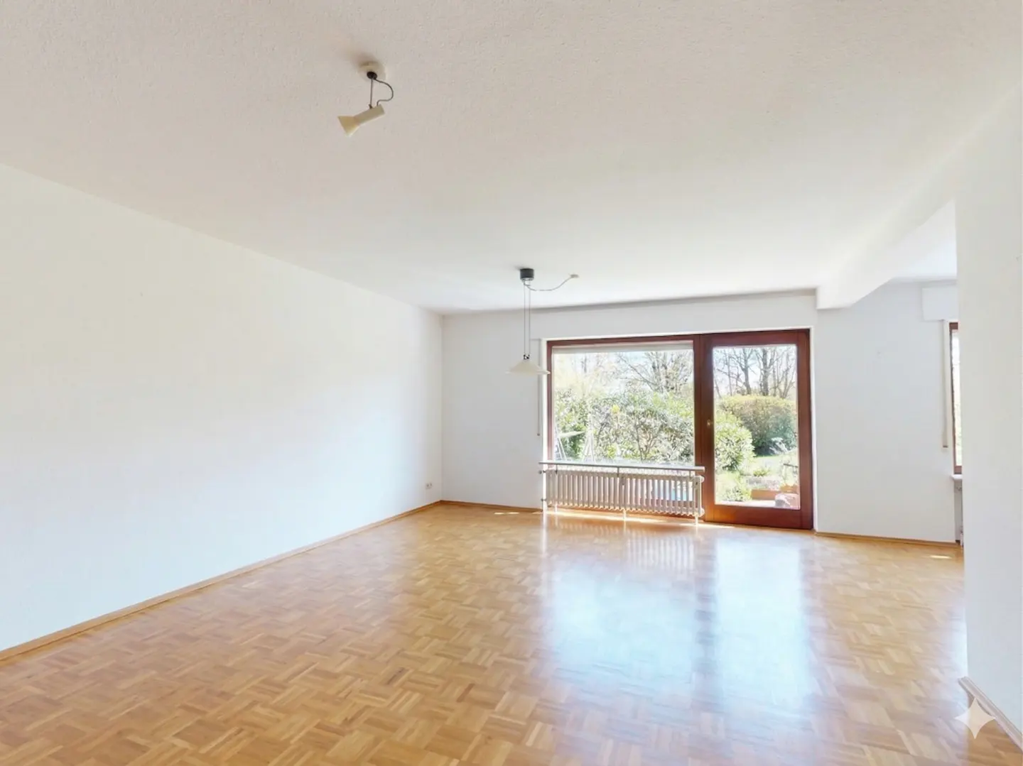 Bright, empty room with parquet floors, white walls, and a large window overlooking a green garden. A radiator sits beneath the window.