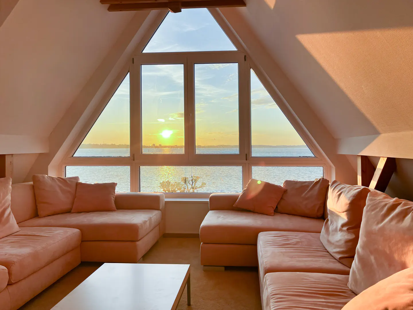 Attic living room with peach sofas, white walls, and a large A-frame window overlooking the ocean at sunset.