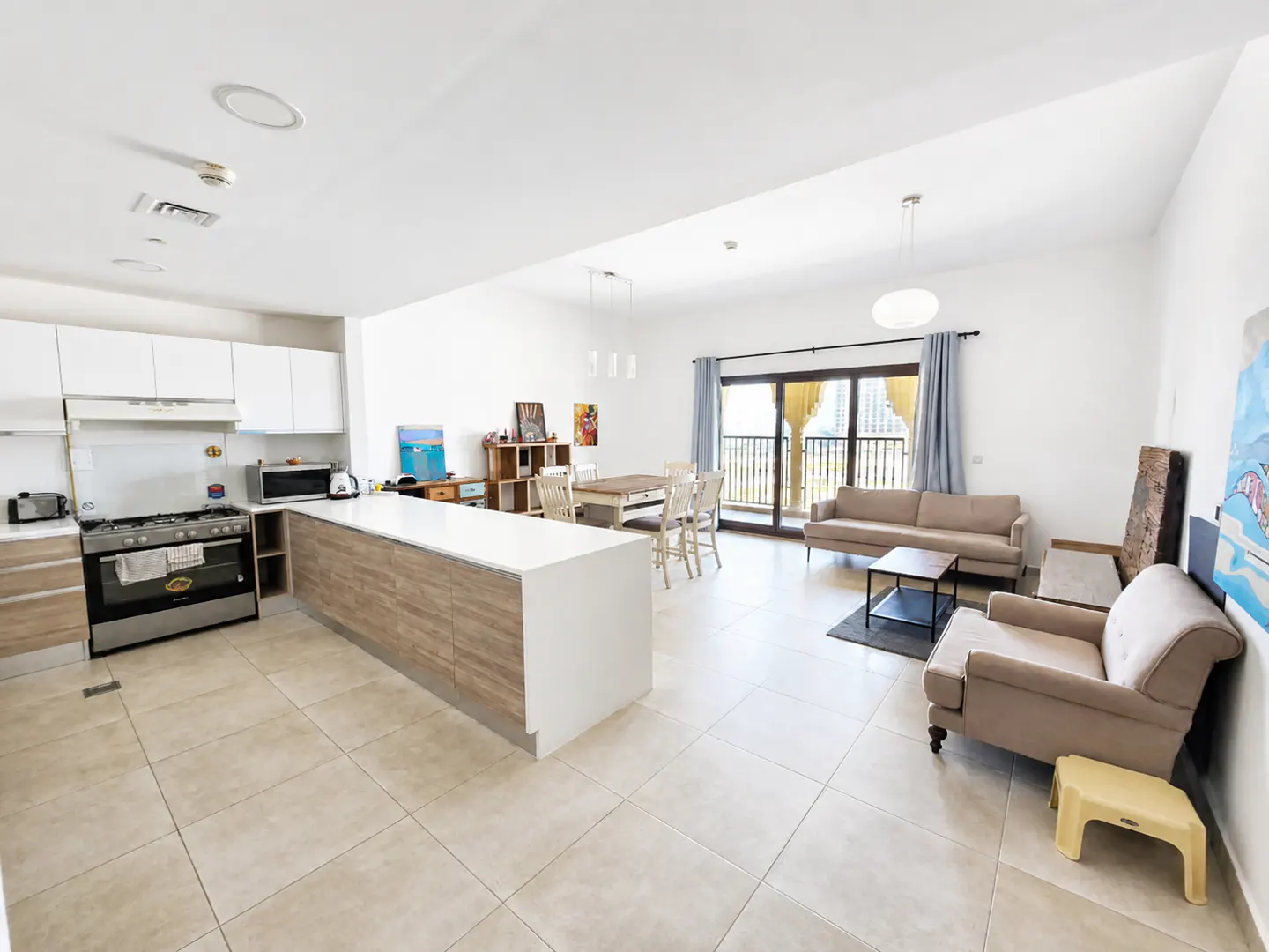 Bright, open-concept apartment with a kitchen island, dining table, and living area with beige sofa and armchair. Balcony with city views.