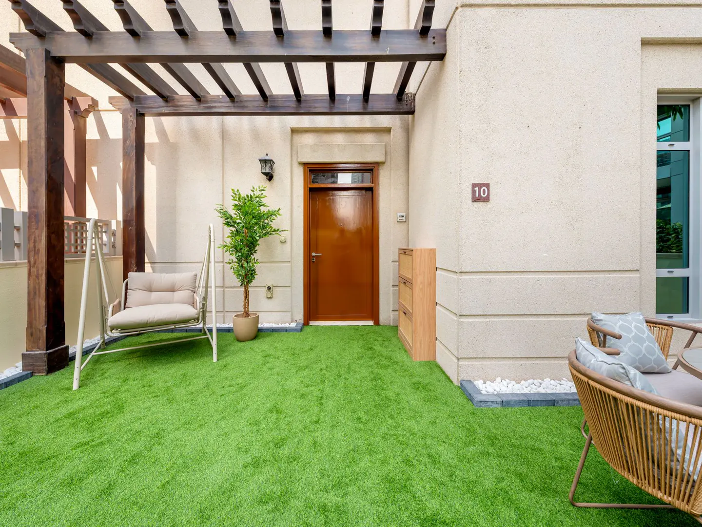 Outdoor patio with green turf, swing, and seating area. Brown door with number 10 on the wall. Wooden pergola above.