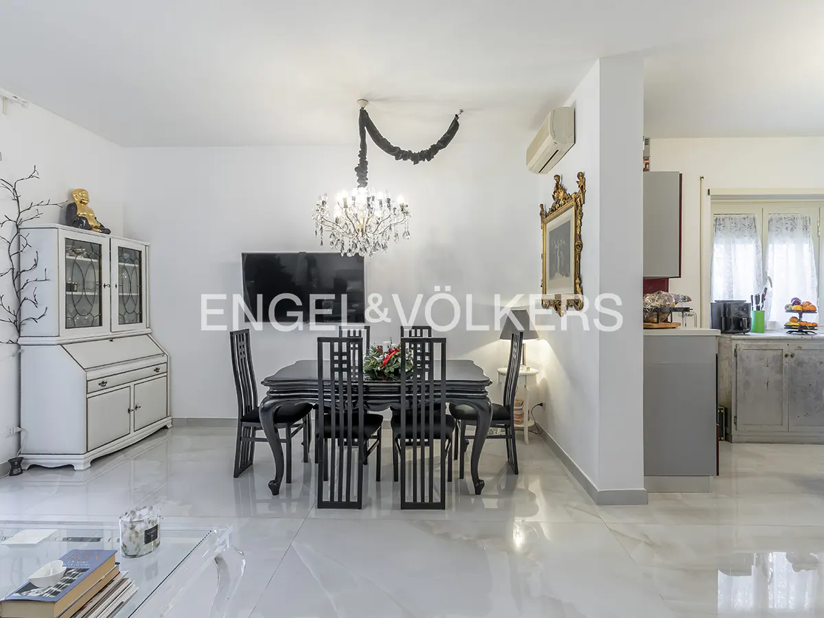 Bright, modern dining room with black table and chairs under a crystal chandelier. White walls, marble floors, and a glimpse of the kitchen.
