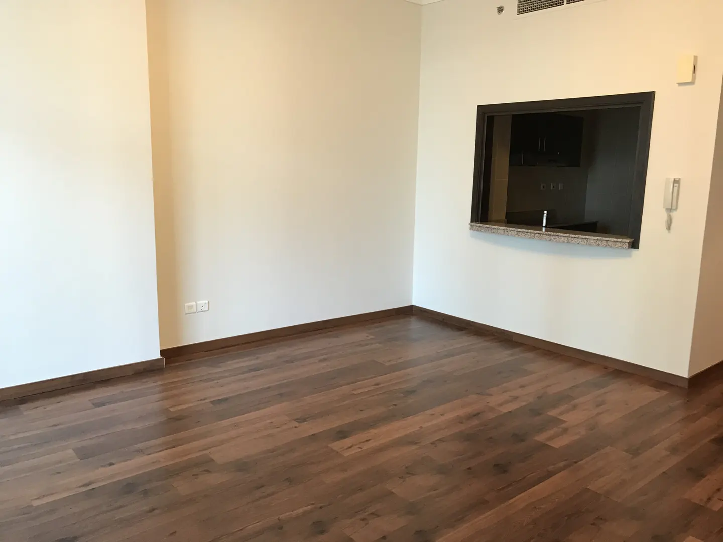 Empty room with dark wood floors and white walls. A pass-through window to the kitchen is visible.