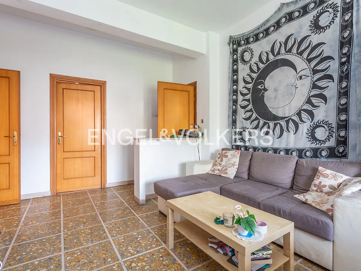 Living room with a gray sofa, light wood coffee table, and a tapestry with a sun and moon design on the wall. Two wooden doors are visible.