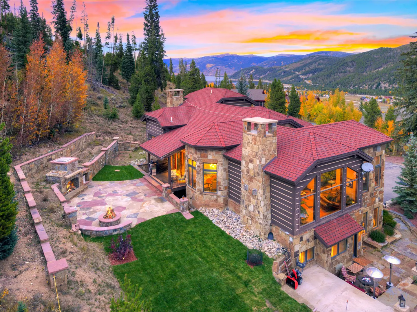 Aerial view of a large stone and wood home with a red roof, patio with fire pit, and mountain views at sunset.