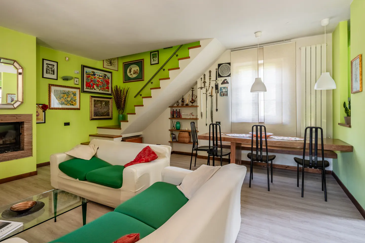 A living room with green walls, white sofas with green cushions, and a wooden dining table with black chairs. Stairs lead to the upper floor.