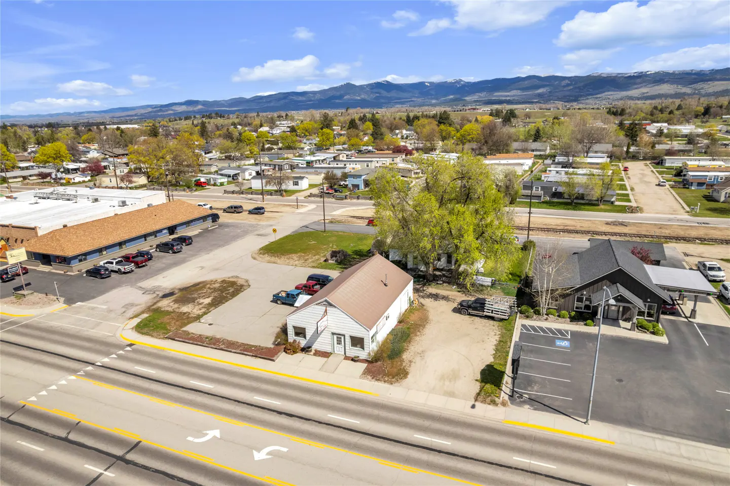 Aerial view of commercial buildings with parking lots, a street with yellow lines, and mountains in the background.