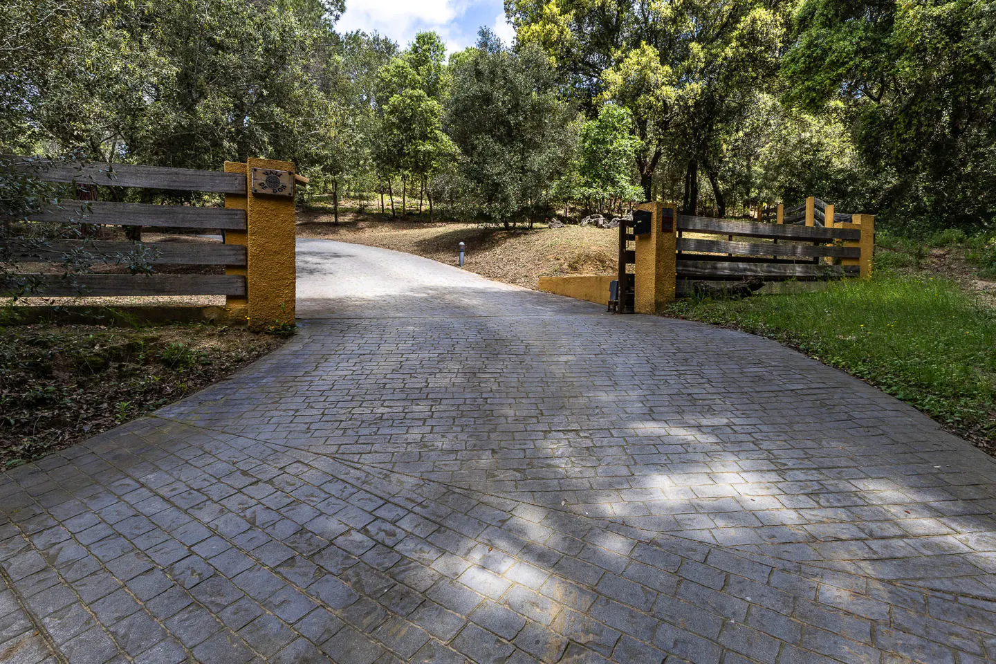 Gated driveway leading to a wooded property. The gate is wood with yellow pillars, and the driveway is paved with gray bricks.