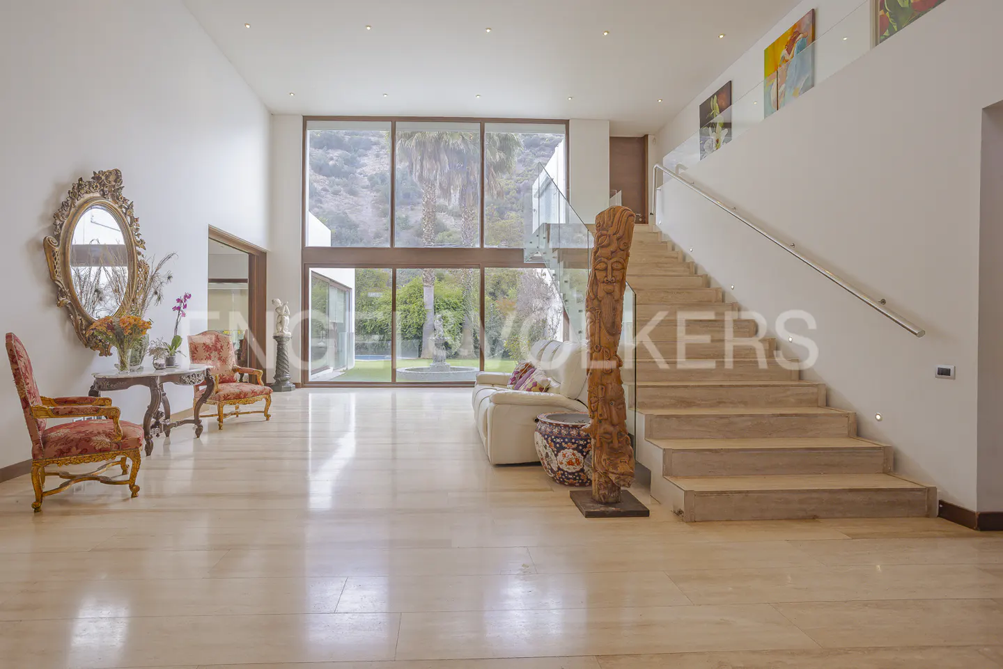 Bright, open foyer with light wood floors, a staircase, and large windows overlooking a green landscape. Ornate mirror and chairs add a touch of elegance.
