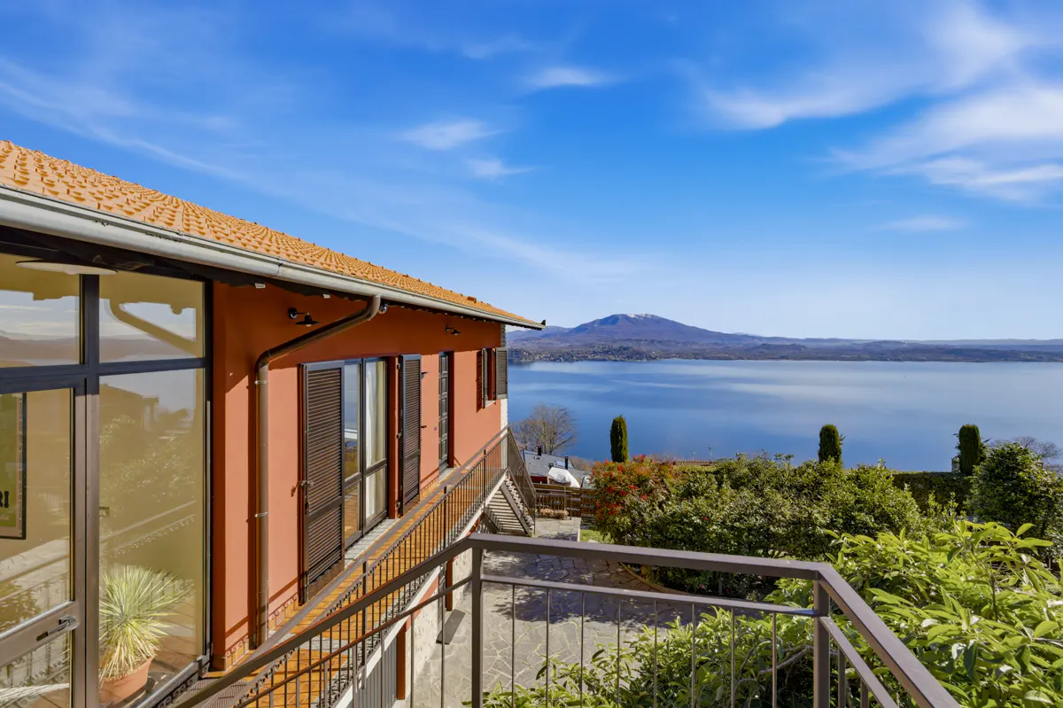 Terracotta-colored house exterior with a balcony overlooking a vast blue lake and distant mountains under a clear sky.