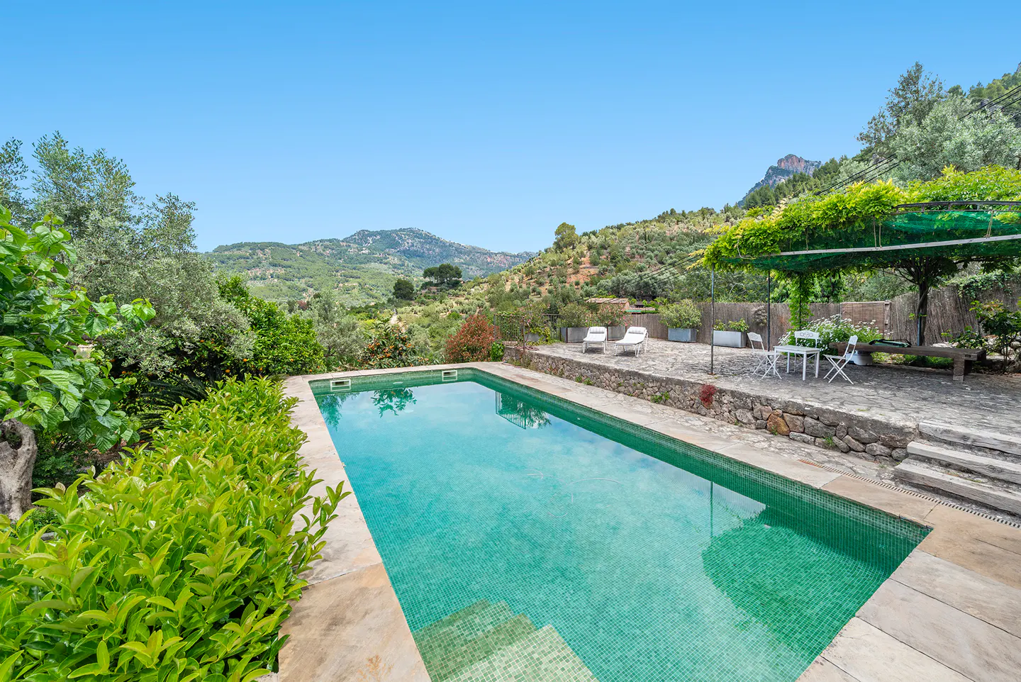 Rectangular pool with turquoise water, stone patio, white chairs, and mountain views under a clear blue sky.