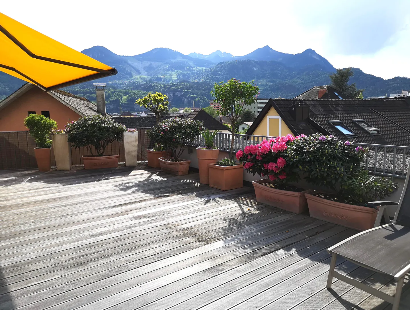 A wooden deck with potted plants and a yellow awning overlooks a town and mountains.