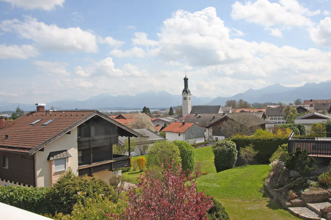 View of a European village with houses, a church steeple, and mountains in the background under a blue sky with white clouds.