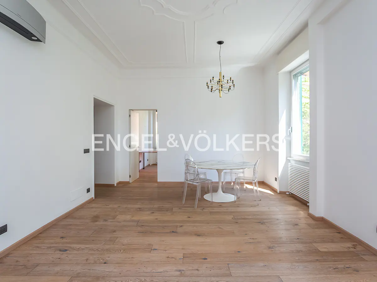 Bright, empty room with hardwood floors, white walls, and a white table with clear chairs. A chandelier hangs from the ceiling.
