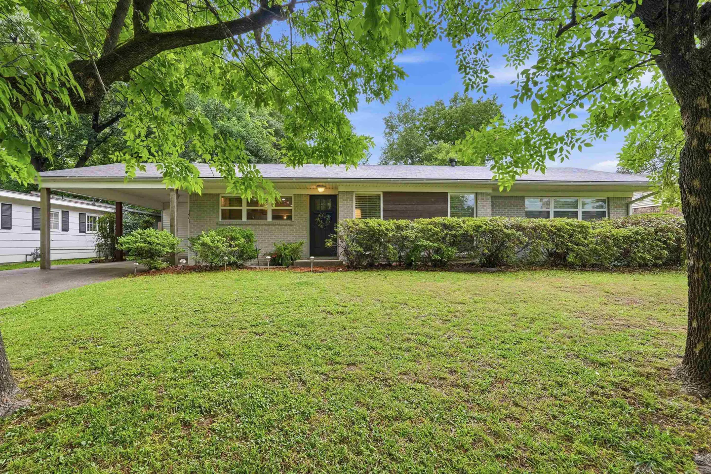 Single-story brick house with a carport, green lawn, and trimmed hedges. Trees frame the view.