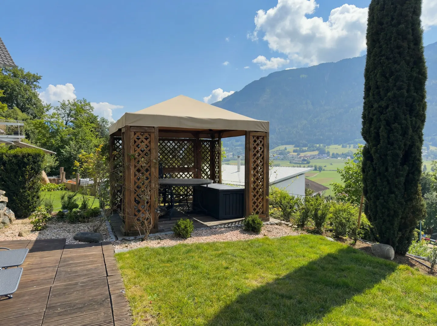 Wooden gazebo with a beige canvas roof, table, and storage box, set in a green lawn with mountains in the background.