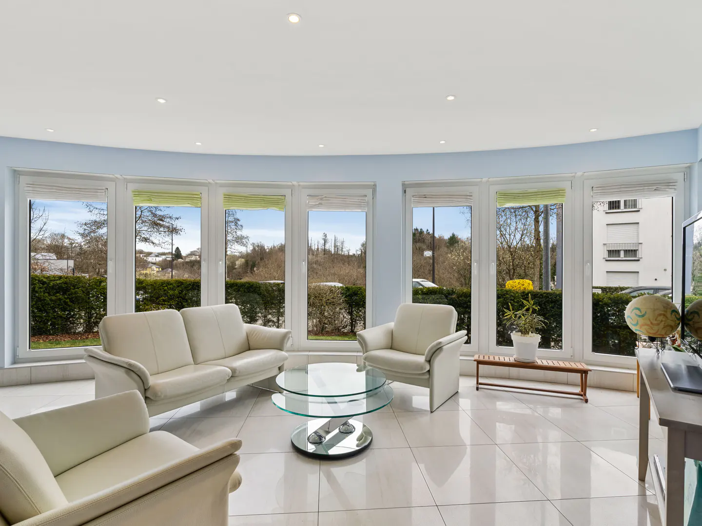 Bright living room with white tile floors, cream sofas and chairs, and a glass coffee table in front of large windows.