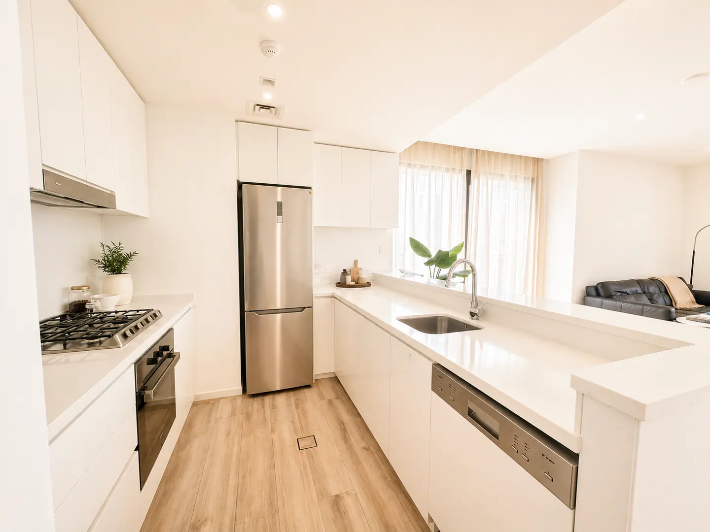 Bright, modern kitchen with white cabinets, stainless steel appliances, and light wood floors. Open layout leads to living area with black sofa.