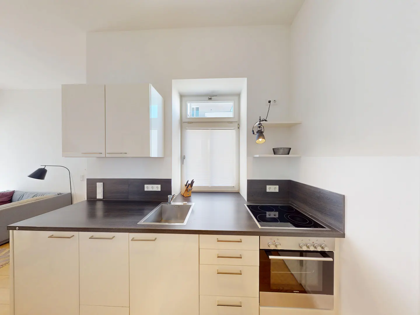 Bright kitchen with white cabinets, dark countertops, and stainless steel sink and oven. A window with a white shade is centered above the sink.