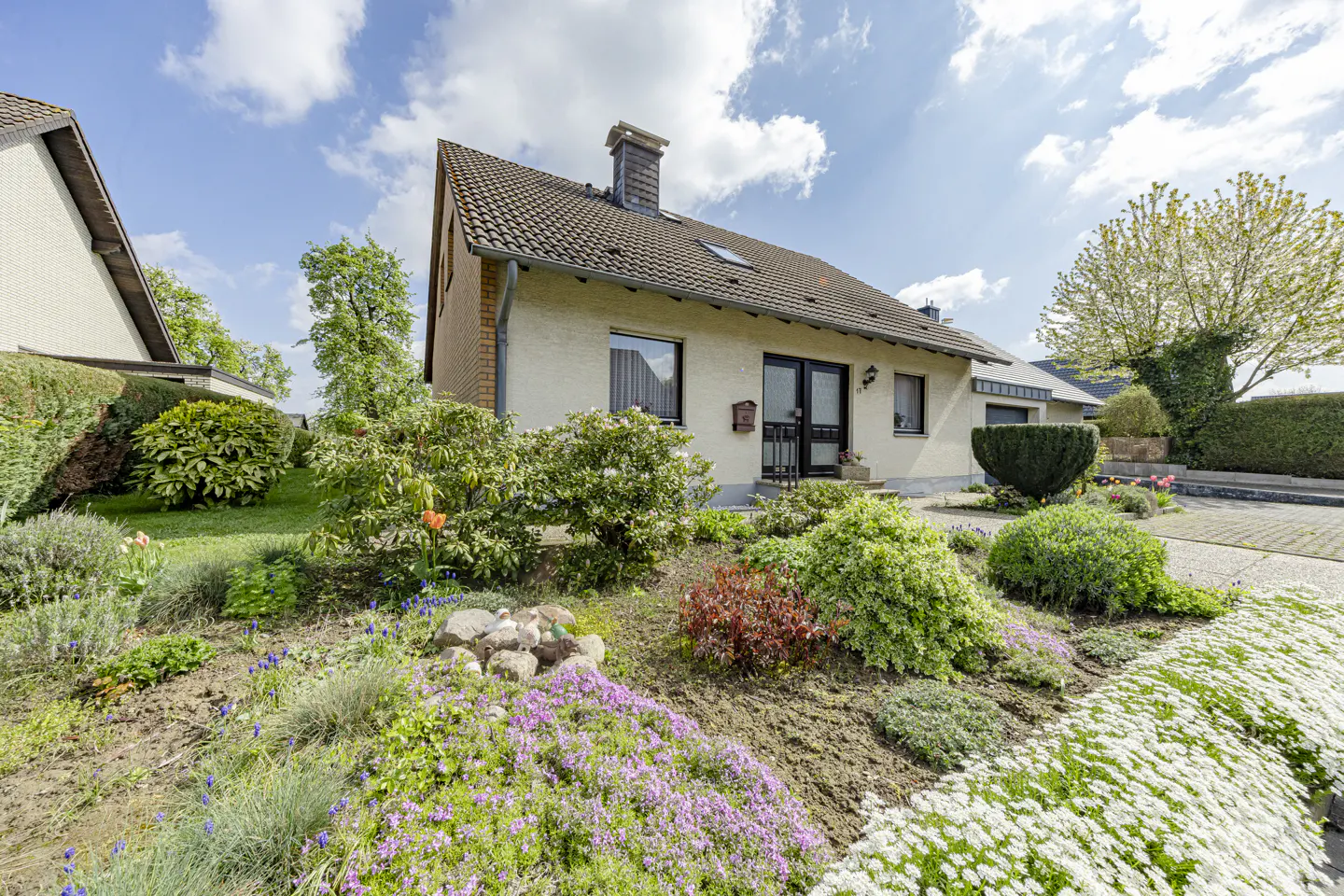 A one-story house with a brown roof, beige walls, and a chimney, surrounded by a lush garden with colorful flowers.