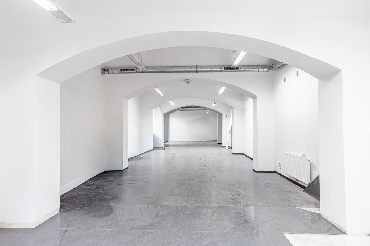 Empty, bright white commercial space with arched ceilings, grey tiled floors, and exposed industrial ventilation pipes. [AI-generated text]