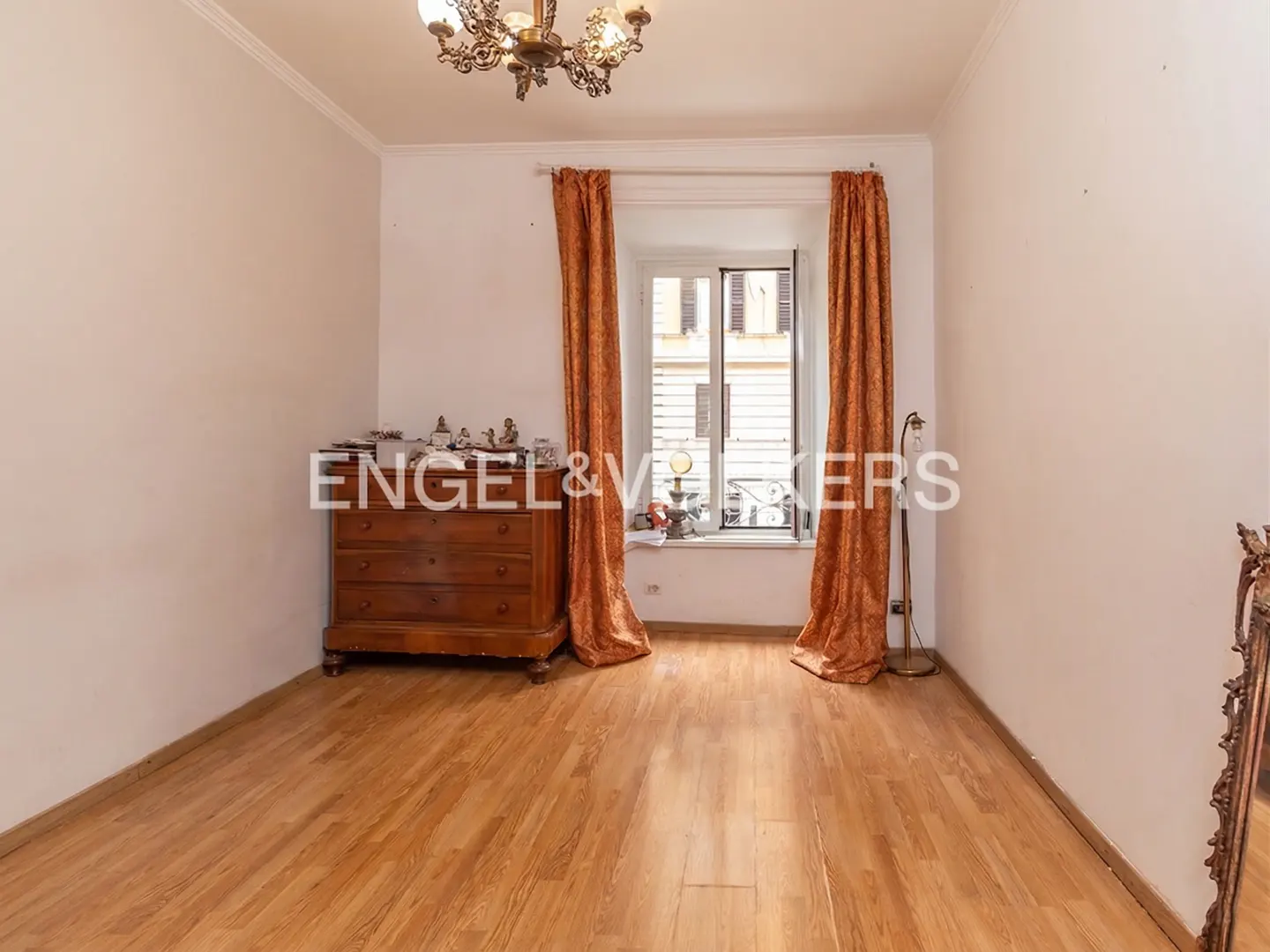 A bright room with hardwood floors, a wooden dresser, and orange curtains framing a window. A chandelier hangs from the ceiling.