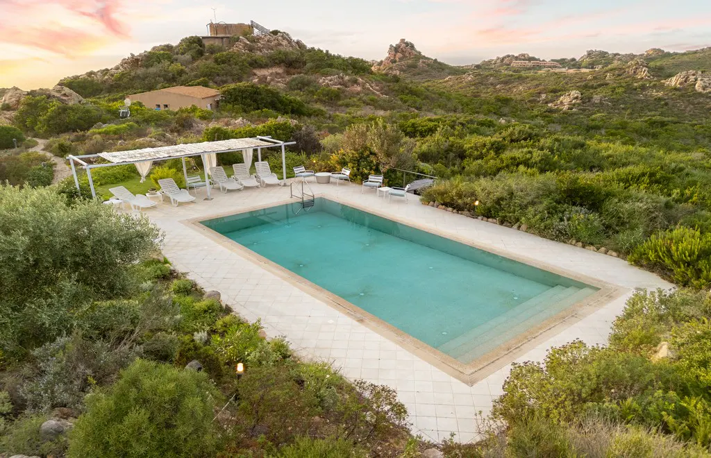 Aerial view of a rectangular pool with turquoise water, surrounded by greenery and lounge chairs, with a building on a hill in the background.