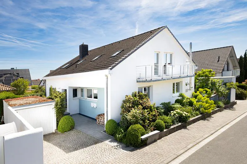 A white two-story house with a brown roof, a balcony, and a well-manicured garden.