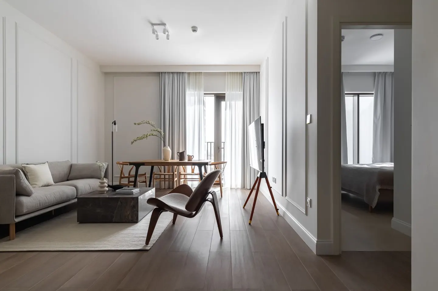 A modern living room with a gray sofa, marble coffee table, wooden dining set, and a TV on a tripod stand. A bedroom is visible through a doorway.