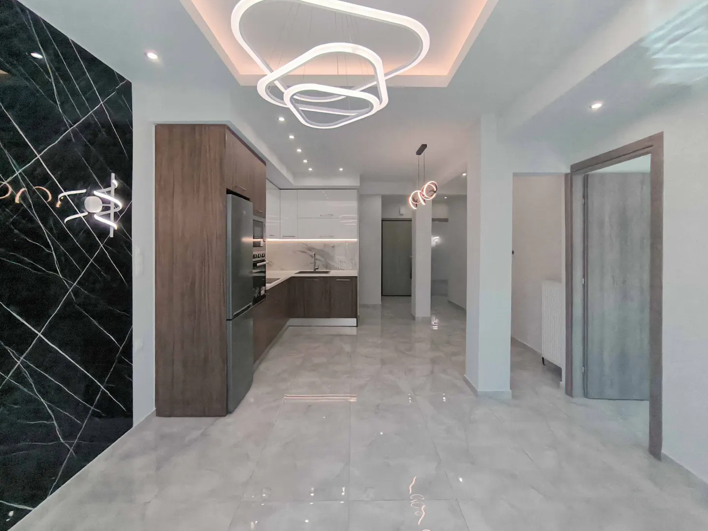 A modern apartment interior with marble floors, a black marble accent wall, and a kitchen with wood cabinets. A unique, white light fixture hangs from the ceiling.