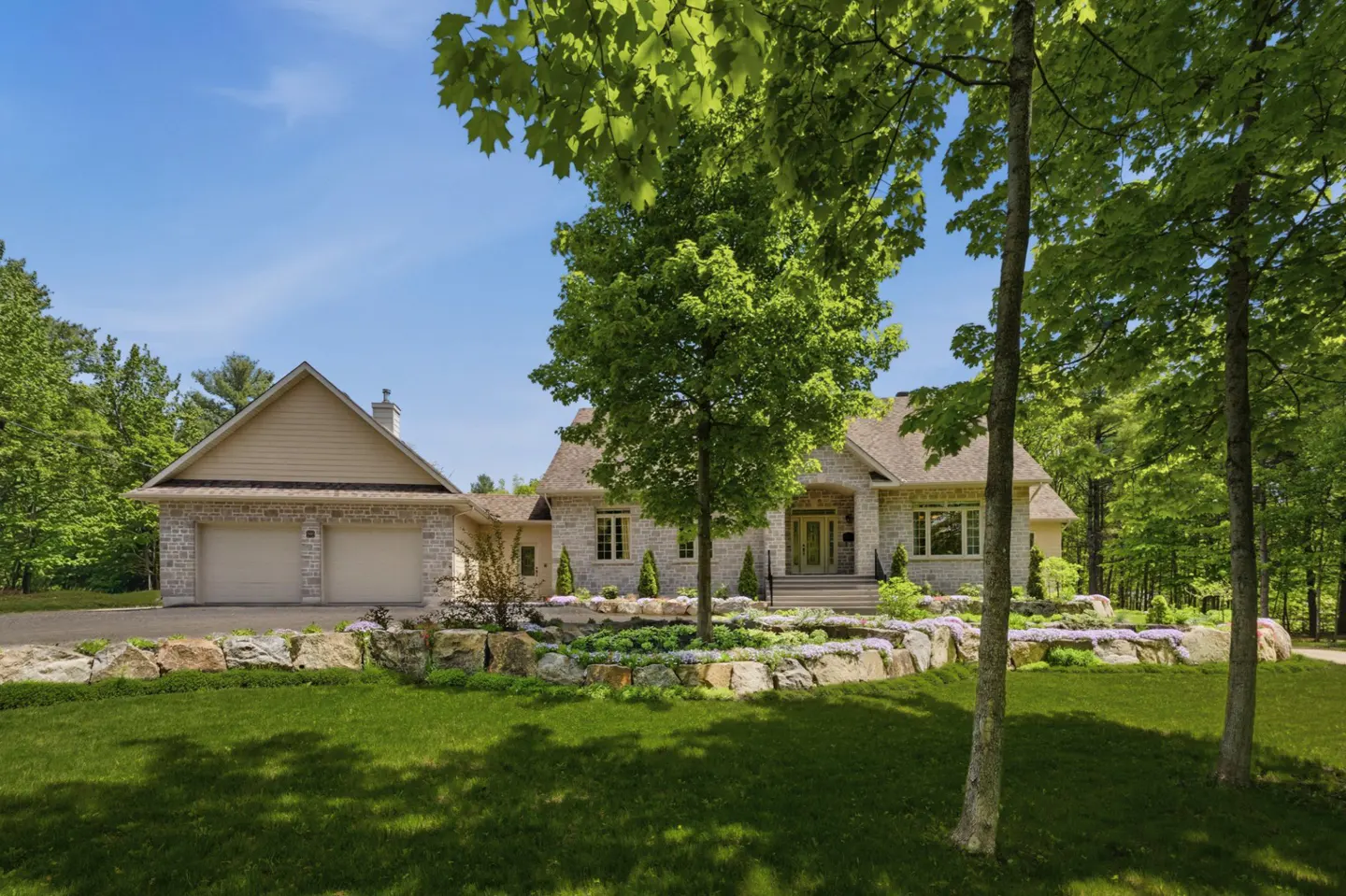 A stone house with a two-car garage, surrounded by lush green trees, a manicured lawn, and a tiered rock garden.