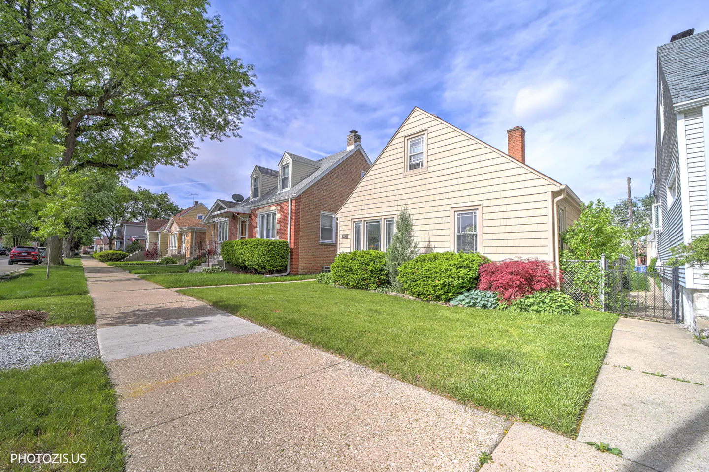 A suburban street view with a sidewalk, green lawns, and various single-family homes under a partly cloudy sky.