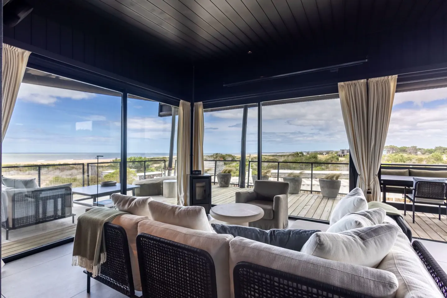 Living room with black ceiling, large windows, and a view of the beach. Neutral-toned sofa, chairs, and curtains create a calming atmosphere.