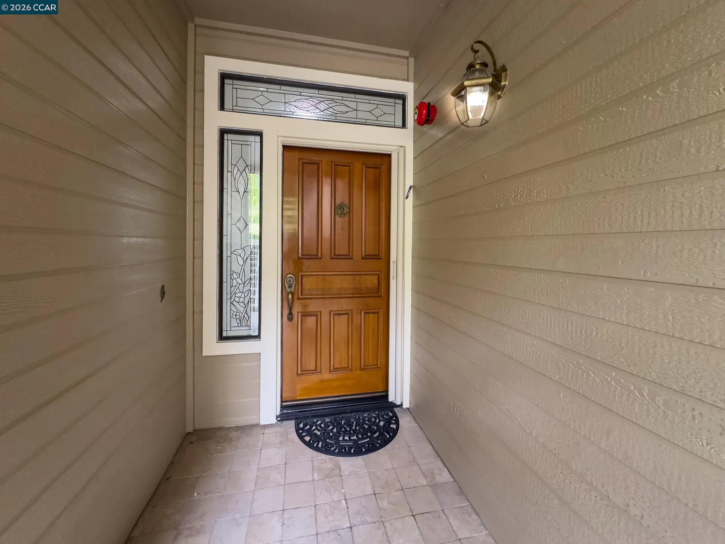 A tan house with a wooden front door, sidelight, transom window, and a black doormat. A brass outdoor light fixture is on the right.