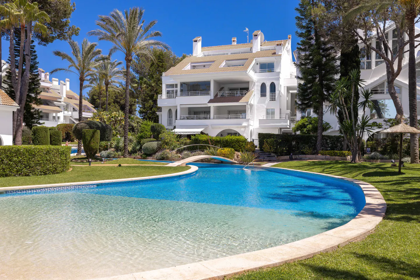 A white multi-story building with balconies overlooks a blue swimming pool and green lawn on a sunny day. Palm trees and other greenery surround the property.