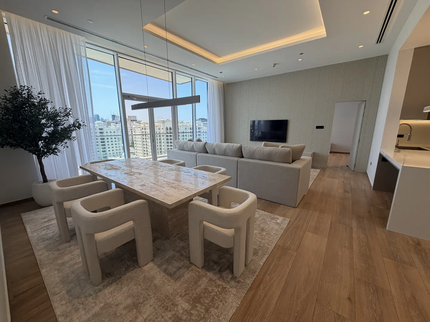 A modern apartment interior with a stone table, white chairs, and a large sofa, with a city view through a large window.