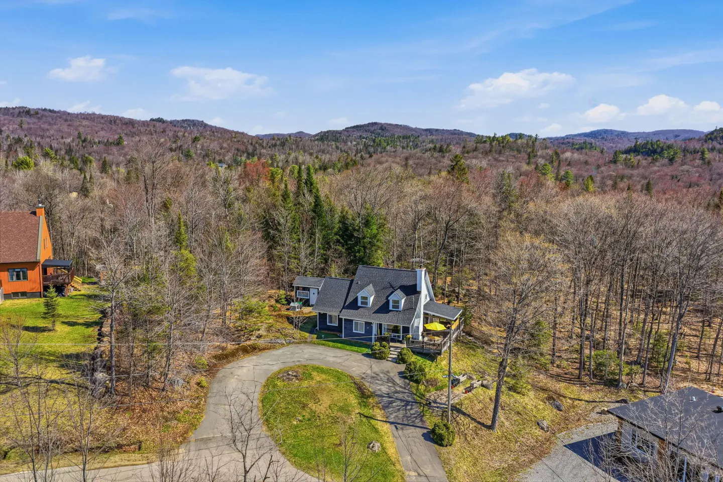 Aerial view of a gray house with a black roof, surrounded by trees and mountains under a blue sky. A circular driveway leads to the house.